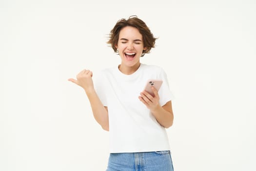 Portrait of beautiful, modern girl with smartphone, pointing finger left, holding smartphone and laughing, posing over white studio background.