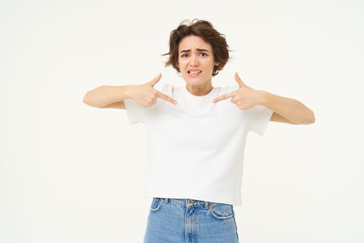 Portrait of worried brunette woman, pointing at herself, looking concerned and anxious, standing over white studio background. Copy space