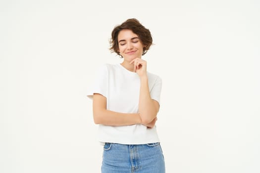 Image of pleased smiling brunette girl, smirks, looks down with confident, satisfied face expression, stands over white background.