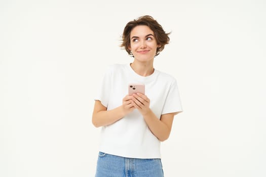 Portrait of cute modern woman with mobile phone, smiling, using smartphone app, doing online shopping on application, standing against white background.