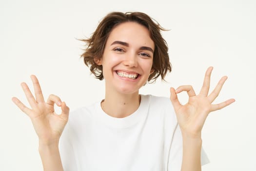 Close up of smiling, happy young woman shows zero, okay ok gesture, approve something good, standing over white background.