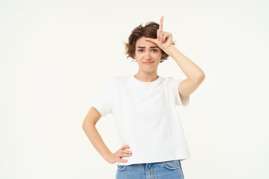 Portrait of arrogant young female model, shows loser, l letter on forehead, stands over white background.
