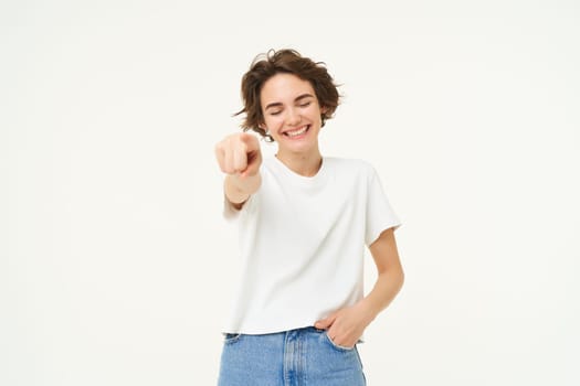 Portrait of excited, smiling young woman, pointing finger at camera, inviting you, congratulating, standing over white studio background.