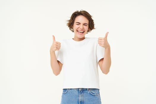 Portrait of cheerful young woman, shows thumbs up in approval, like something good, nod in approval, looking confident, standing over white background.