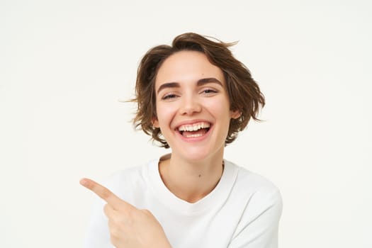 Close up shot of happy, candid woman, laughing and smiling, pointing finger left, showing advertisement, standing over white background.