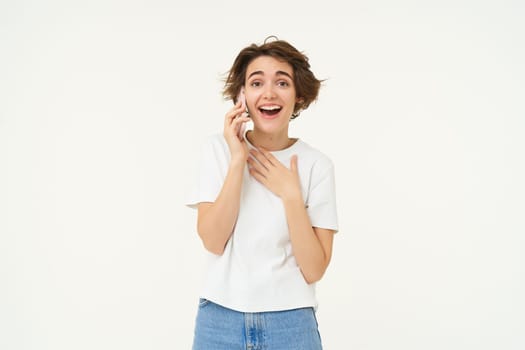 Portrait of chatty young woman talking on mobile phone, laughing and smiling, answer telephone with surprised face, standing over white background.