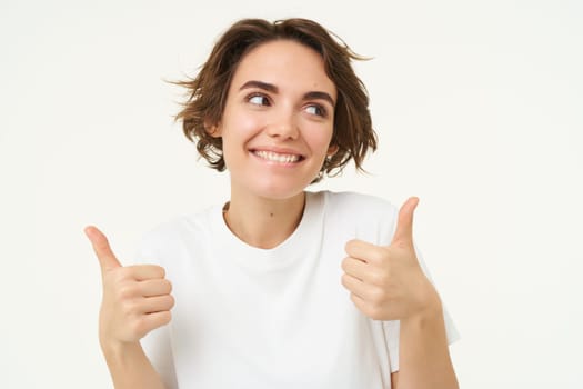 Close up shot of cheerful smiling girl, shows thumbs up, gives approval, likes something, positive feedback concept, stands over white background.