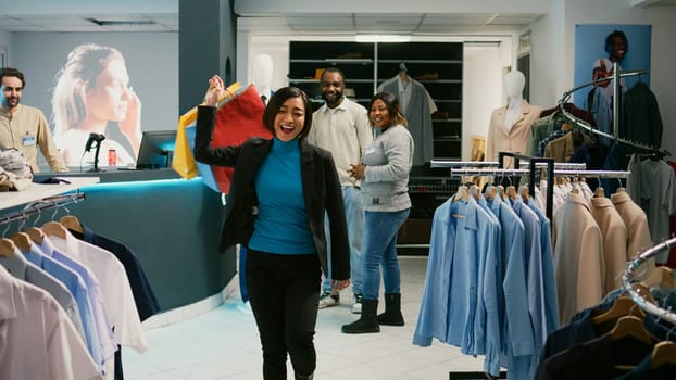 Asian customer doing dance moves with shopping bags, feeling happy after making purchase at cash register. Young woman dancing and buying trendy clothes at shopping mall. Tripod shot.