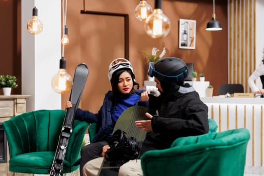 Young couple dressed in winter gear sitting on sofa in lounge area. Man and woman holding their wintersport equipment talking in hotel lobby ready for snow adventure. Indoor winter holiday vibes.