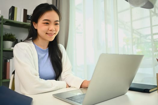 Satisfied Asian female freelancer searching information, working on laptop computer at home.