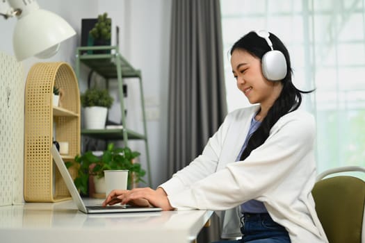 Pleased young woman wearing headphone and using laptop, studying online, typing email or working remotely.