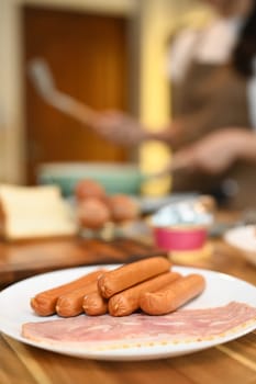 Close up a white plate of sausages and bacon on a wooden kitchen counter for breakfast.