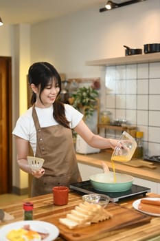 Young woman in apron pouring a beaten eggs from a glass bowl into a frying pan.