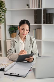 Attractive young businesswoman sitting at desk with documents and using a digital tablet.