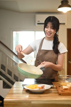 Pretty young woman serving scrambled eggs from a pan onto a white plate for breakfast.