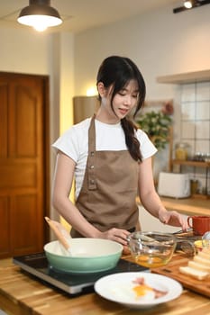 Attractive young asian housewife wearing apron preparing breakfast in the kitchen.