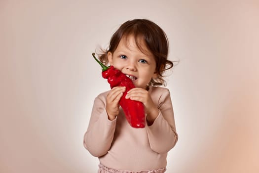 cute baby girl bites fresh red pepper on beige background. healthy baby food. kid loves vegetables
