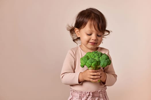 smiling child girl looking at fresh green raw broccoli on beige background. healthy baby food. kid loves vegetables