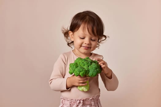little child girl holding fresh green raw broccoli on beige background. healthy baby food. kid loves vegetables