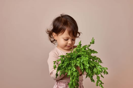 adorable child girl holding fresh green parsley on beige background. healthy baby food.