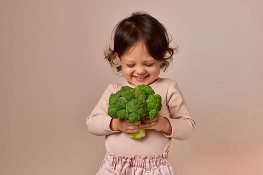 smiling cute child girl holding fresh green raw broccoli on beige background. healthy baby food. kid loves vegetables