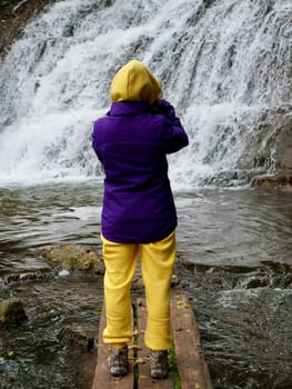 A woman stands with her back to the river in autumn in a yellow suit and purple jacket. The person travels through the Ukrainian Carpathians to the Chervonohorod Dzhuryn waterfall. Vertical