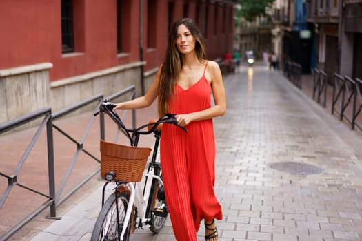 A fashionable woman in a vibrant red dress strolls with her ebike along a charming cobblestone street, surrounded by elegant architecture, enjoying the relaxed atmosphere of the city