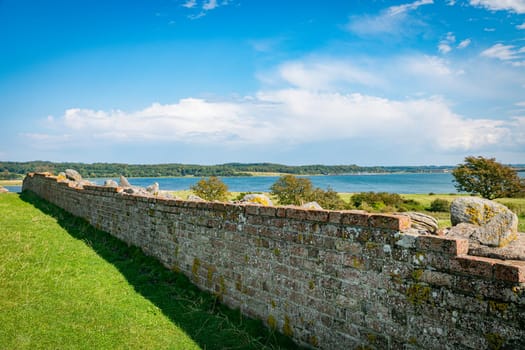 at kalo ruine in denmark at mols bjerge, A scenic view of a historic stone wall stretching along a lush green field, with a tranquil lake and forested hills in the background under a bright blue sky.