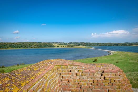Beautiful lakeside scene featuring an old, moss-covered brick wall in the foreground with lush greenery and a clear blue sky at mols bjerge jutland denmark