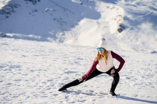 Full body of young female in warm clothes and sunglasses, standing on snowy slope in winter while practicing yoga with stretched legs and hands on knee warrior pose