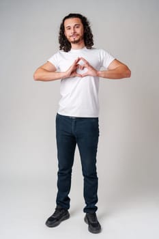 Young man with curly hair forms a heart shape with his hands while standing against a light background in studio