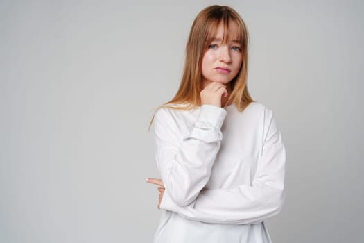 Young woman in a white long-sleeve shirt thoughtfully poses with a hand on her chin in a minimalistic indoor setting