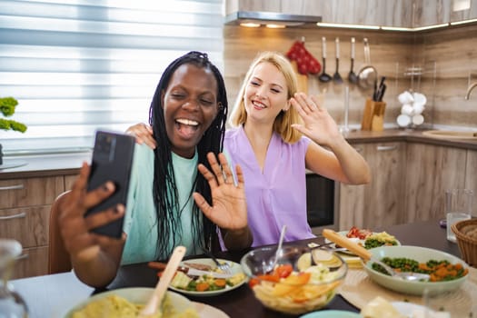 Two friends enjoy a delightful meal while smiling and waving at a device.