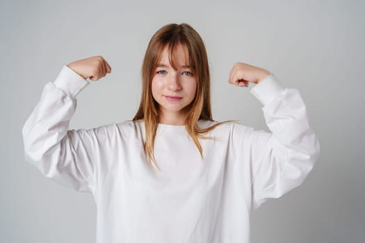 A joyful young girl with long hair wearing a white sweater stands against a plain background close up