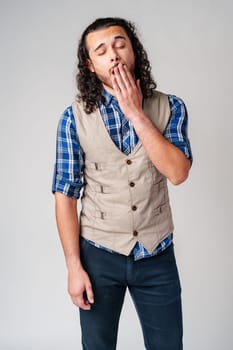 A young man with curly hair yawning while wearing a plaid shirt and vest poses in a neutral studio setting