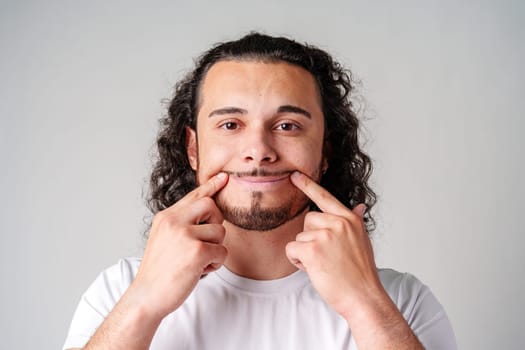 Young man in a white t-shirt playfully smiles while pointing to his teeth against a plain gray background in a studio setting
