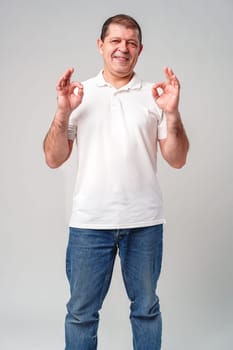 A man in a white shirt poses with a cheerful expression showing OK hand gestures in a well-lit indoor setting close up