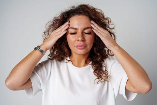 Woman with curly hair holds her head in discomfort, expressing concern or stress in a neutral backdrop close up