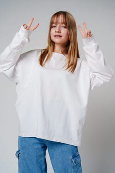 Young woman joyfully posing with peace signs against a plain background in a casual white sweatshirt