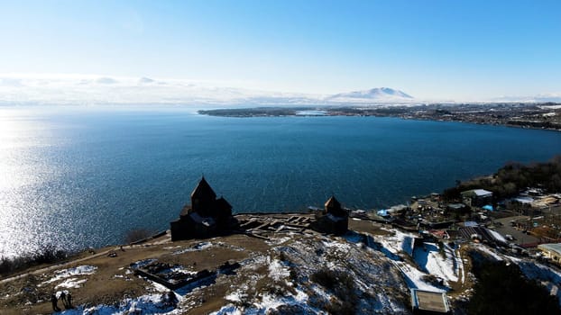 Aerial view of a hill with wooden old buildings. Footage. Early spring with hills covered by snow and calm blue sea with clear sky above