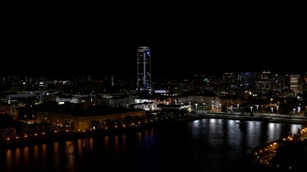 The view from the drone.Stock footage.A beautiful city at night located next to the river and beautiful high-rise new buildings and offices and with a calm illuminated road. High quality 4k footage