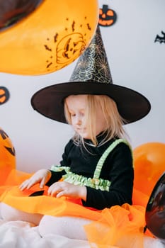 Children's Halloween - a girl in a witch hat and a carnival costume with airy orange and black balloons at home. Ready to celebrate Halloween.