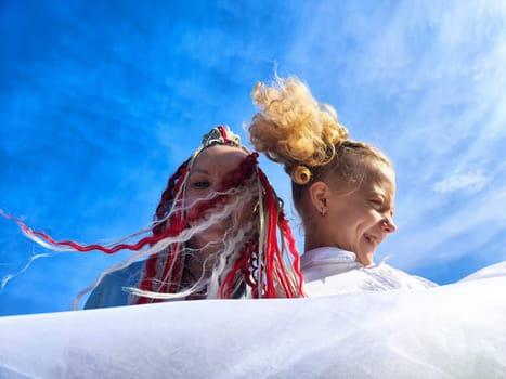 The joyful atmosphere features two women laughing and showcasing their vibrant hairstyles under a clear, sunny sky.