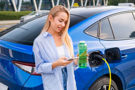 Smiling woman using her phone while charging an electric car and drinking water from the green bottle. Eco friendly and renewable energy concept.