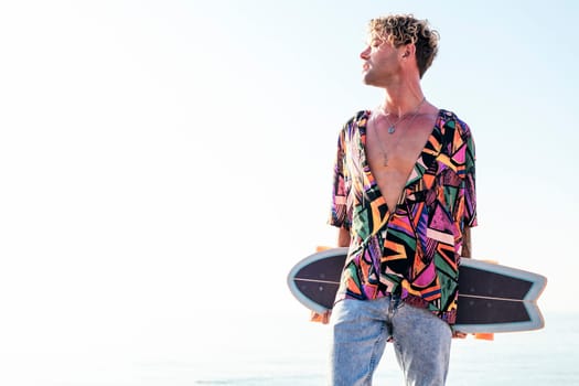 young hipster male skater in trendy summer outfit carrying skateboard while standing on embankment against sea and cloudless sky in summer day