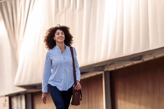 satisfied ethnic businesswoman with bag walking along street and smiling while looking forward