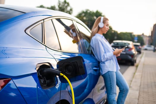 Close up electric car charging by the roadside, while a woman stands nearby wearing headphones, listening to music