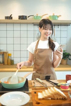 Smiling young woman using smartphone while preparing meal on a wooden worktop.
