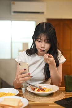 Beautiful asian woman having breakfast and using smartphone at kitchen.
