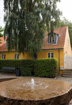 ebeltoft,denmark,A quaint yellow house with a red-tiled roof stands amidst lush greenery. A rustic stone fountain in the foreground adds a tranquil, serene atmosphere to the picturesque scene